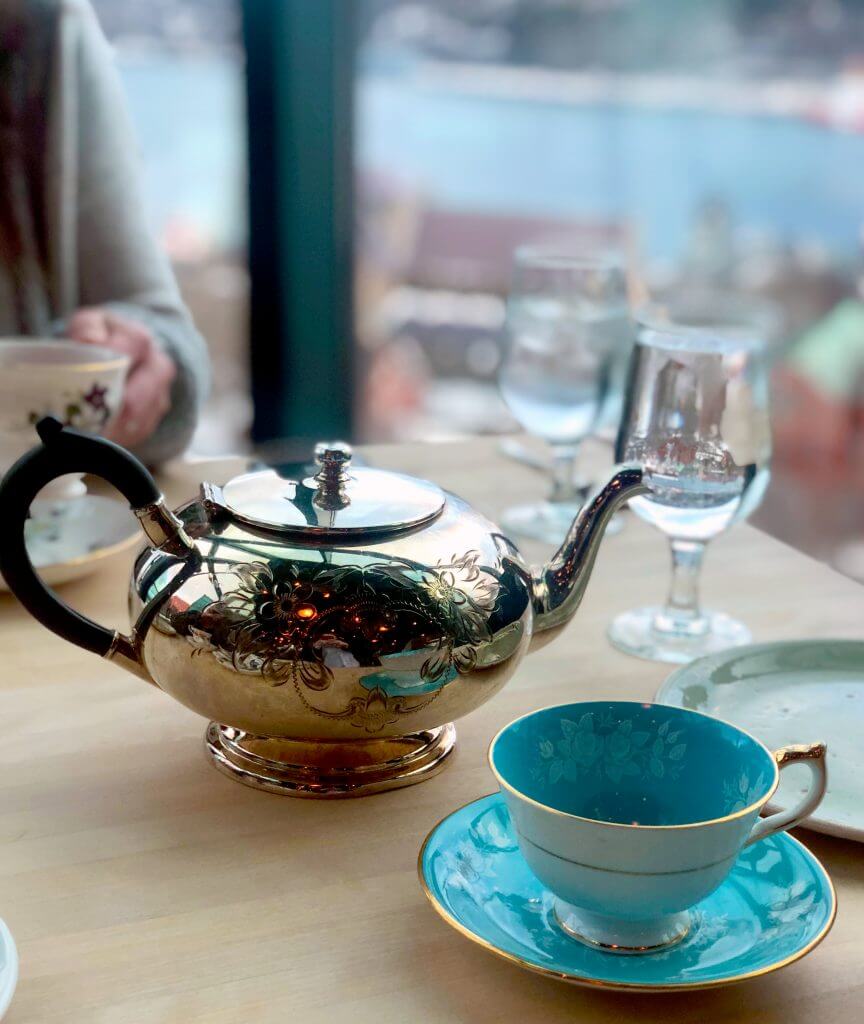 A close up shot of a silver tea pot next to a beautiful blue cup and saucer set with a light flower pattern and gold trim. In the background a guest holds a white tea cup.