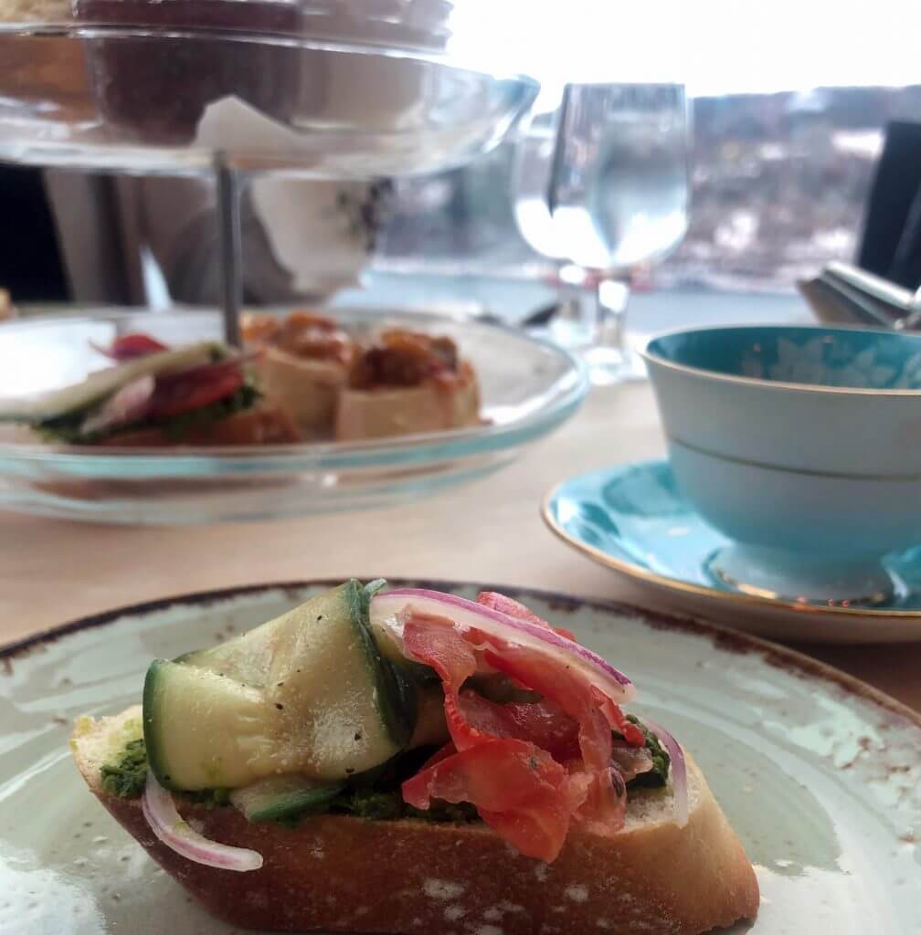 In the foreground, an open-faced vegetarian sandwich on a crostini. In the background there is a cup and saucer set as well as a serving stand full of treats.