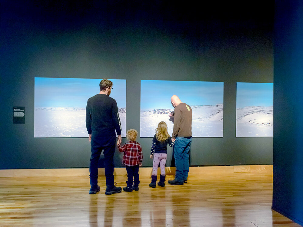 Two children holding their dads' hands as they look at art on a wall in The Rooms.