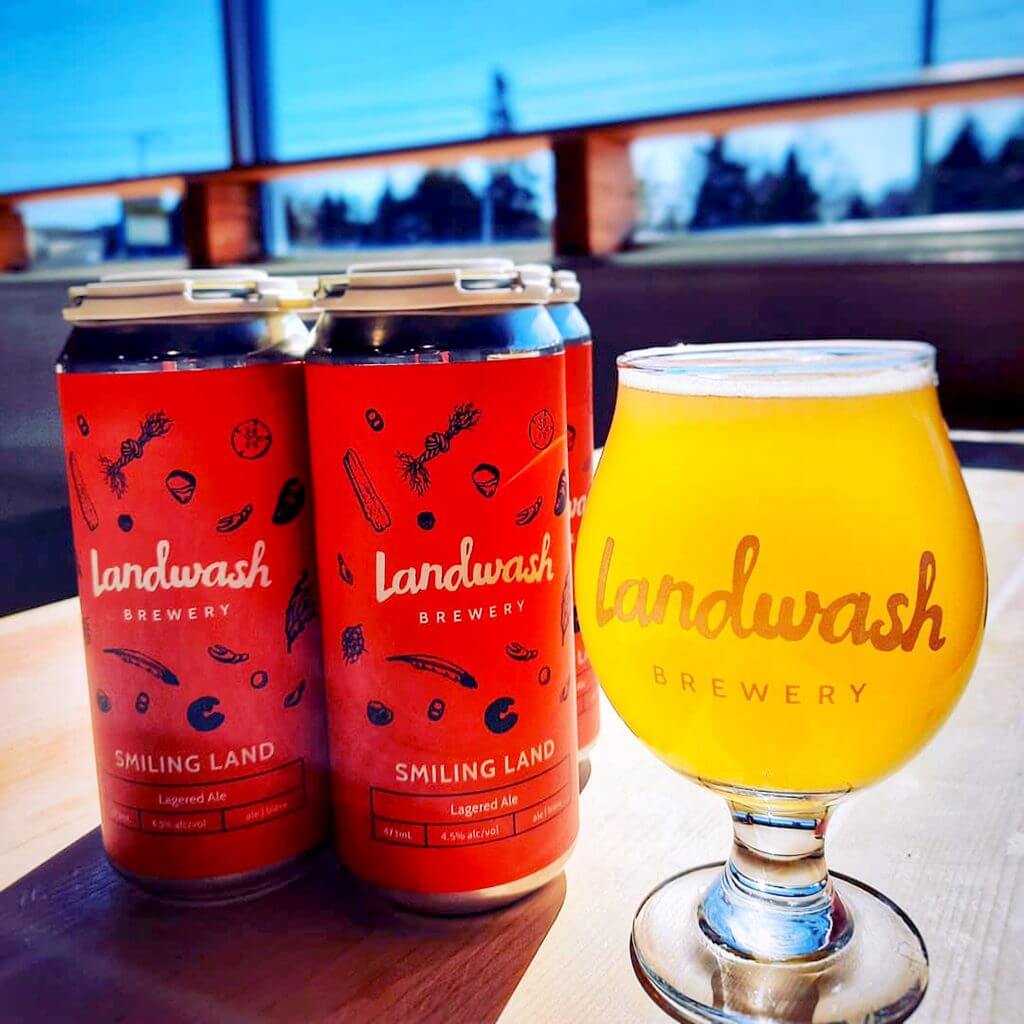 A glass full of craft beer sits on a table next to two cans of Landwash Smiling Land.