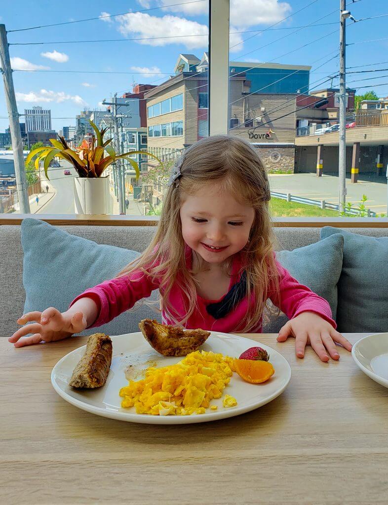 Blake with a big smile on her face looking lovingly at a plate of eggs, sausages, and fruit.
