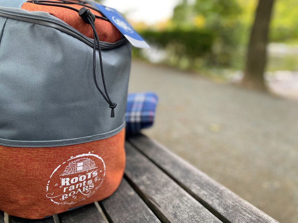 A Roots Rants Roars backpack in grey and orange sits on a park bench with a blue picnic blanket tucked behind it.