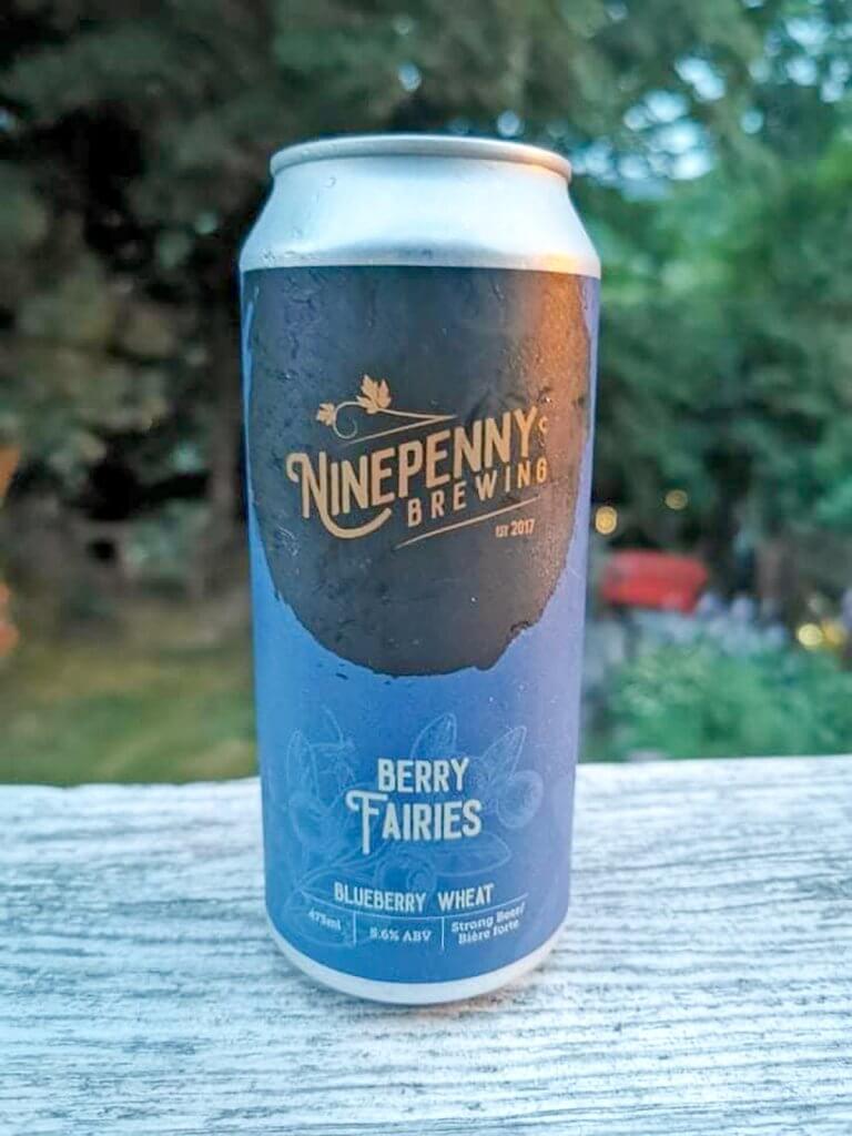 A can of Ninepenny's Berry Fairies sits outdoors on a piece of wood with a backyard in the background. 