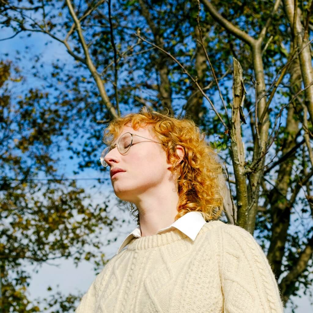 Kelly wearing a white cable knit sweater outdoors with trees in behind her. She looks wistful.