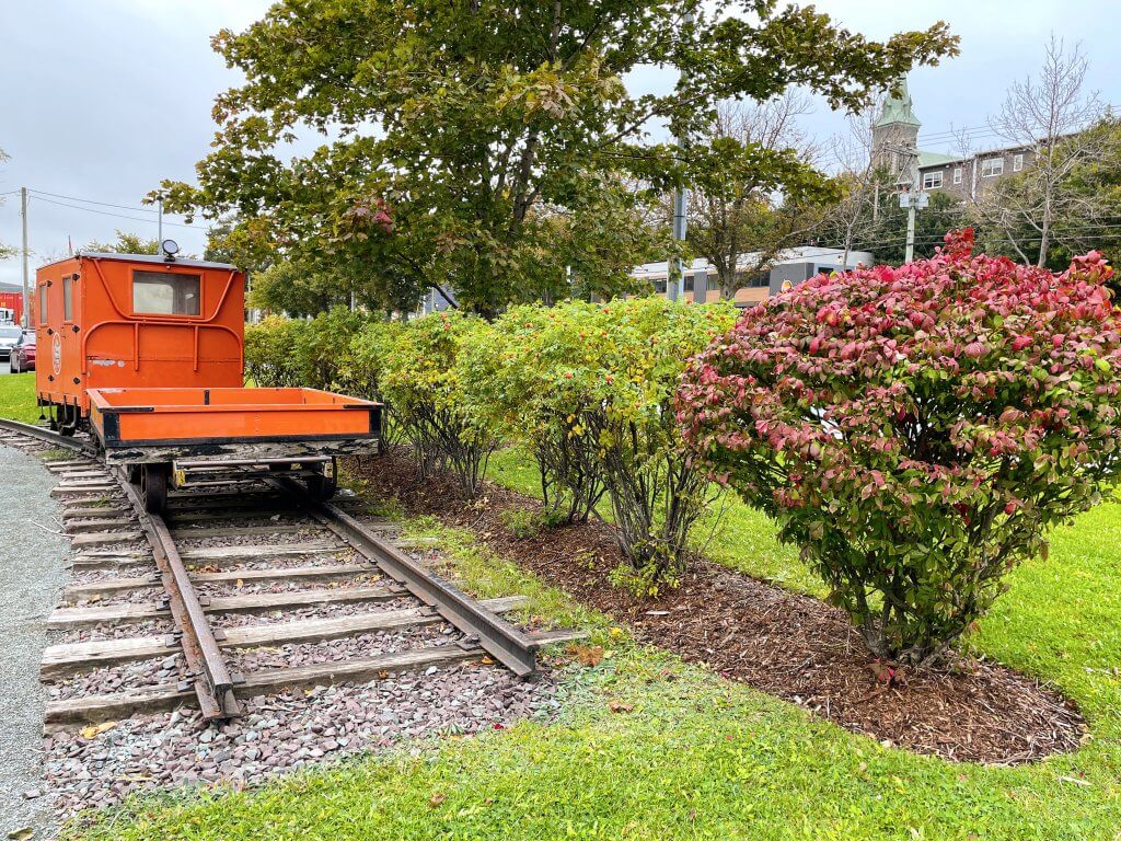 An orange train car on a track with trees all around. Gorgeous fall colours are present.
