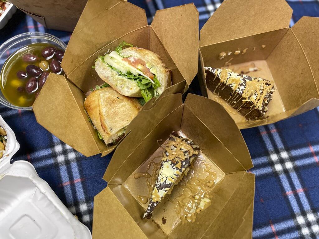 Three cardboard containers sitting on a picnic blanket. One has sandwiches. The other two each have a piece of delicious looking chocolate torta.