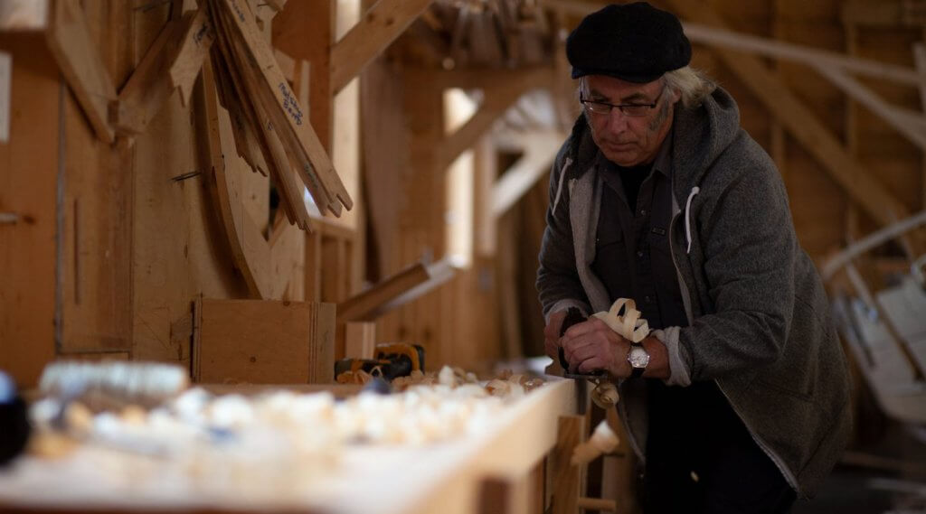 A man wearing a traditional cap planes a wooden boat at the Wooden Boat Museum in Winterton, Newfoundland & Labrador