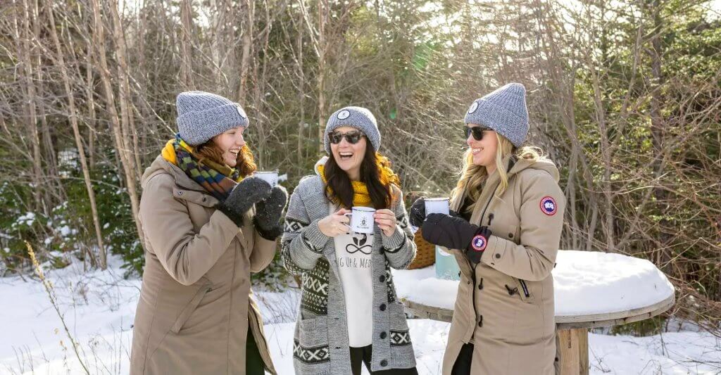 Three warmly-dressed women stand to enjoy a hot mug up in the woods during winter time on Newfoundland