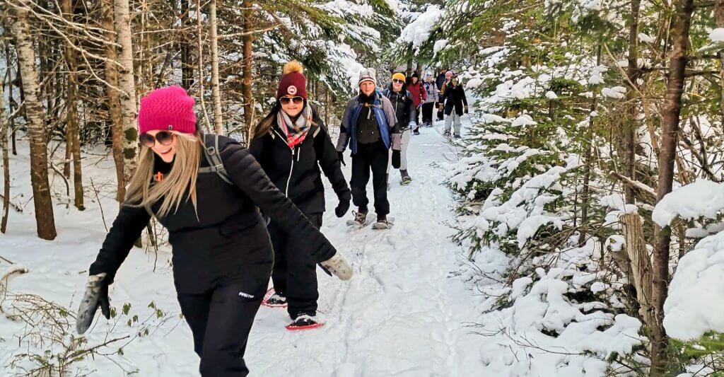 Several woman are seen snowshoeing in single file on a groomed snowy path through the woods on Newfoundland